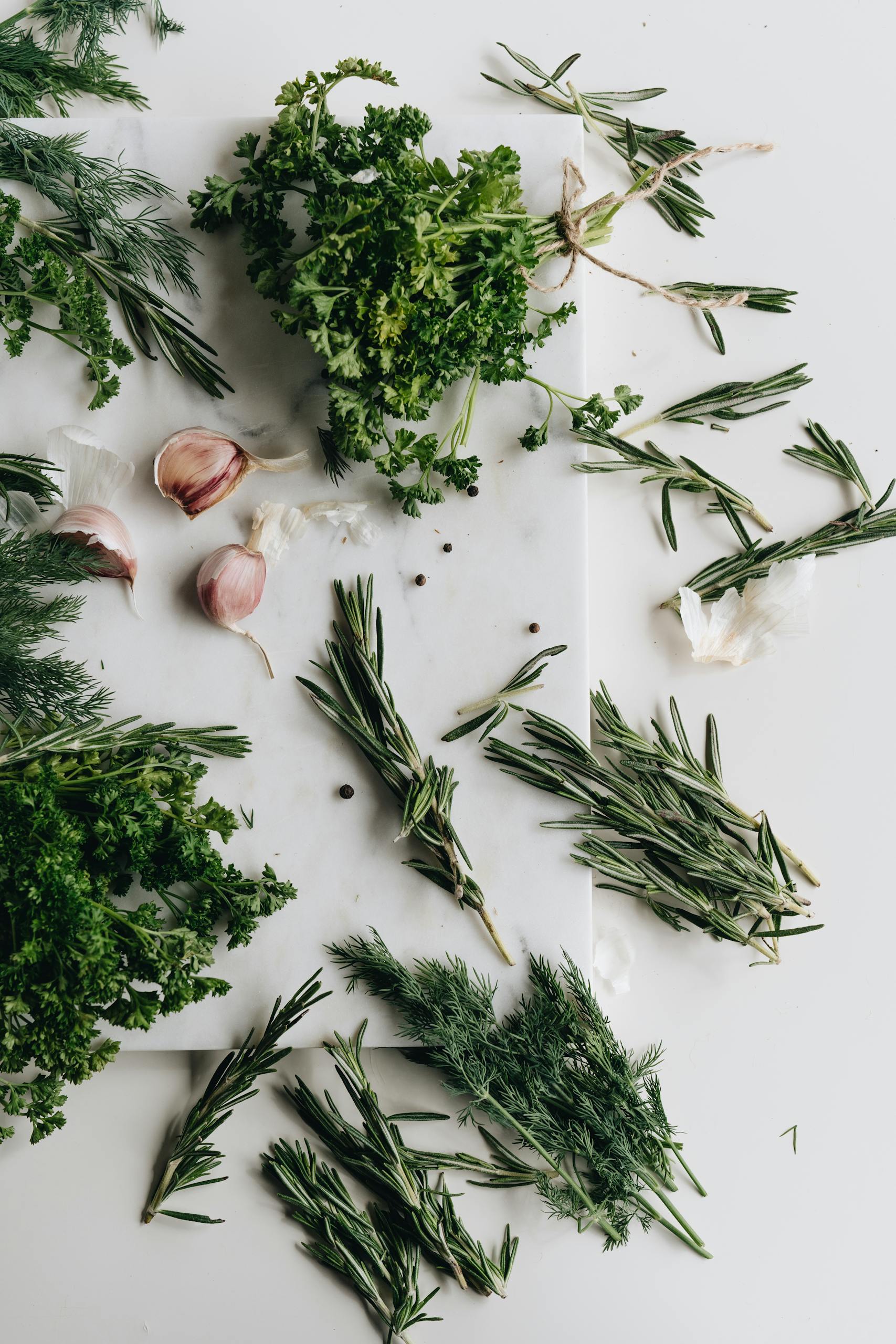 A variety of fresh herbs including garlic, parsley, and rosemary on a rustic white table, perfect for culinary use.
